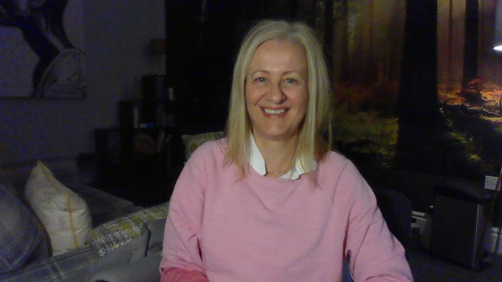 A woman with long blond hair, a pink sweatshirt and smiling at her office where individual appointments happen at Counselling West Bridgford, Nottingham. About me