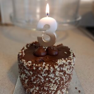 Chocolate cake with cream sprinkles on the side, candle shaped as number 3 on top, candle is lit