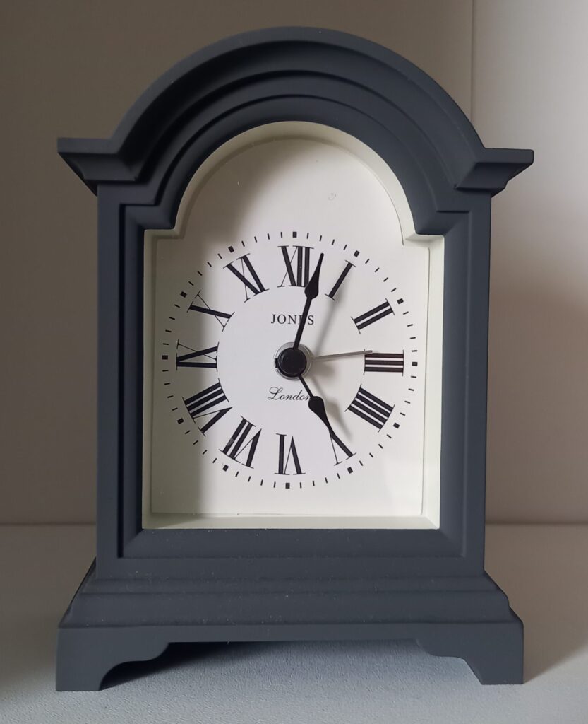 Small grey carriage clock with a grey face and roman numerals for numbers. For Drop-in appointment page.