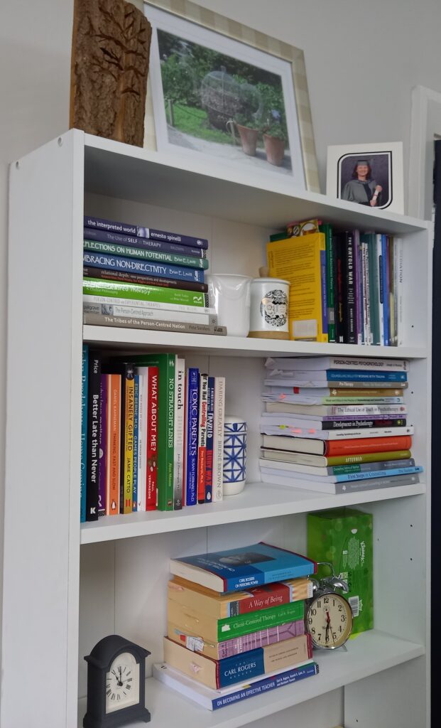Book case with piles of books about psychotherapy and psychology, 2 clocks, a photograph of Michelle at her graduation, a framed photo of a garden, a wooden ornament with the shape of a pine tree cut out